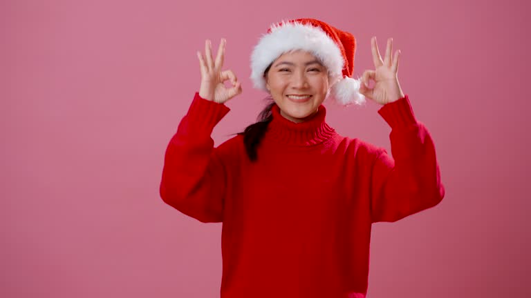 Asian woman wearing Santa hat happy confident showing OK sign by hand standing isolated over pink background.