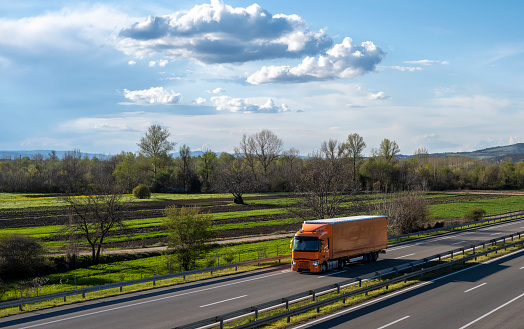Orange semi trailer truck driving on a highway with dramatic sky in the background. Transportation vehicle. Orange truck driving on asphalt road in a rural landscape.