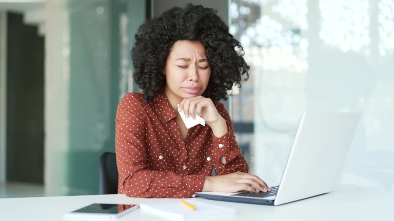 Sad young african american female employee crying and suffering from mental pain sitting at workplace in office.