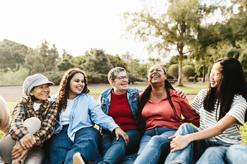 Happy multi generational group of women with different ethnicities having fun sitting on grass in a public park - Females empowerment concept