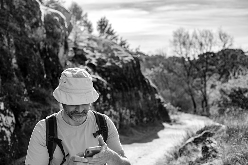 Parque de la cuenca alta del manzanares. Madrid, Spain. Black and white photograph of a hiker with a hat in a natural setting, looking at their mobile phone.