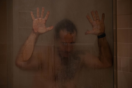 40-year-old man in the shower, resting his hands and face on the steam-fogged shower head.