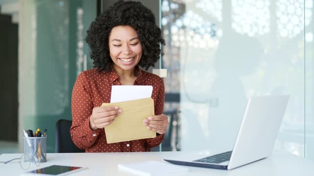 Happy excited young african american female employee reading letter with great news sitting at workplace in office