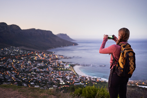 Rear view of a woman hiker taking photo of the city along the coast with her mobile phone while standing on the mountain trail in evening