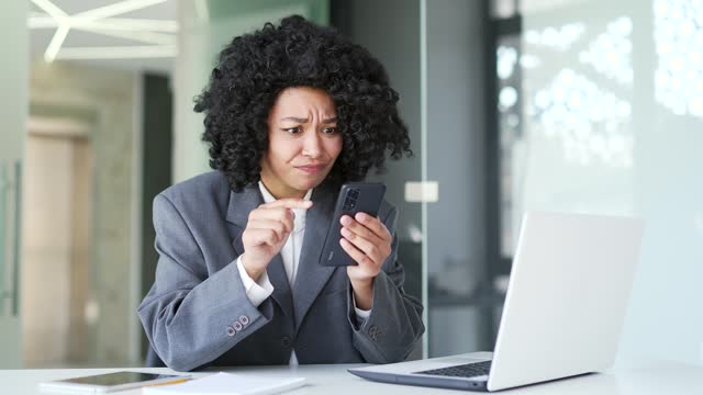 Frustrated young african american businesswoman complains about poor performance of software on smartphone sitting in office.