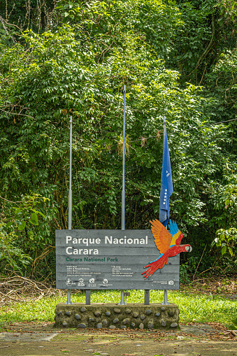 Costa Rica, Parque Nacional Carara - July 22, 2023: Closeup, official white on gray wood welcome sign with image of scarlet macaw and blue flag. Green foliage as backdrop
