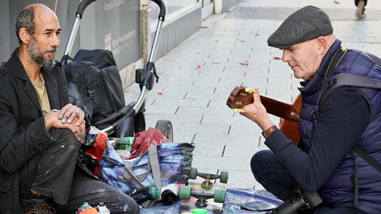 Cardiff, Wales, Oct 29 2023:  A street artist, fingers caked in spray paint, chats with a street photographer who plays the guitar he uses for busking, enjoy each others company and forge a friendship