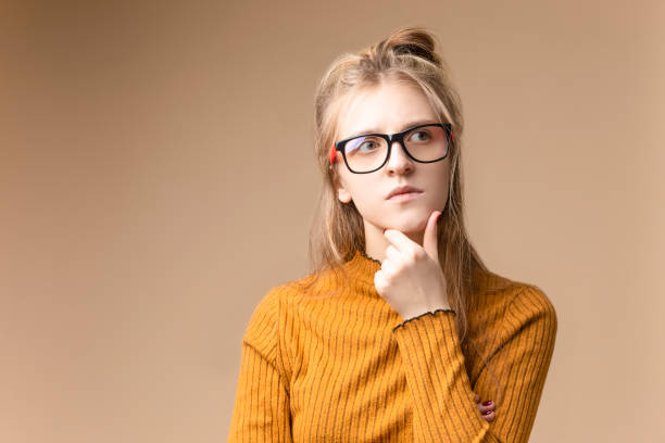 istock Tranquil Thoughtful Caucasian Blond Female in Yellow Knitted Sweater Posing In Glasses While Touching Chin On Beige Background. 1798992952