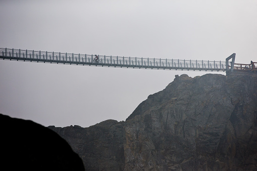 A man rides his mountain bike across the Whistler Cloudraker Skybridge suspension bridge on a cloudy, rainy day. The Whistler Cloudraker Skybridge is located at the Whistler Mountain Bike Park in British Columbia, Canada.