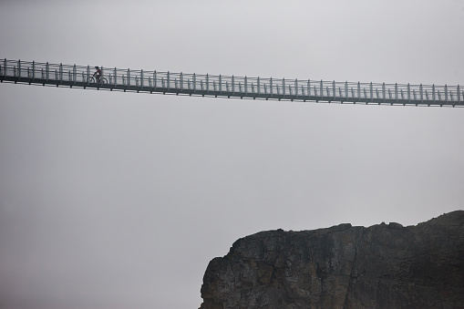 A man rides his mountain bike across the Whistler Cloudraker Skybridge suspension bridge on a cloudy, rainy day. The Whistler Cloudraker Skybridge is located at the Whistler Mountain Bike Park in British Columbia, Canada.