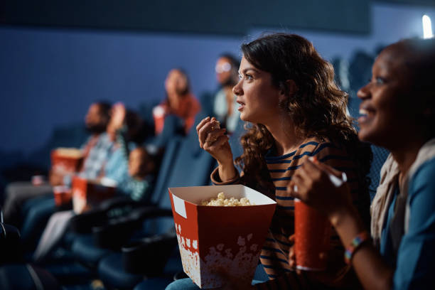 Young woman eating popcorn while watching suspenseful movie in cinema. Young woman watching a movie with anticipation while being with her friends in theater. suspenseful stock pictures, royalty-free photos & images