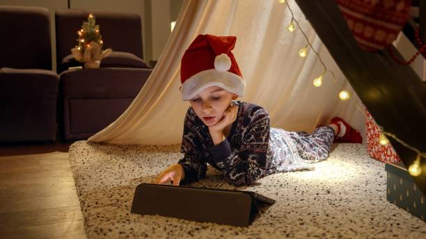 istock Little boy in Santa's hat lying in tepee tent and watching video on tablet computer on Christmas eve. Winter holidays, celebrations and party. 1799059231