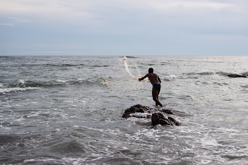 Salvador, Bahia, Brazil - January 29, 2022: Fisherman is seen on top of a rock on the beach of the Red River throwing a fishing net. City of Salvador, Bahia.