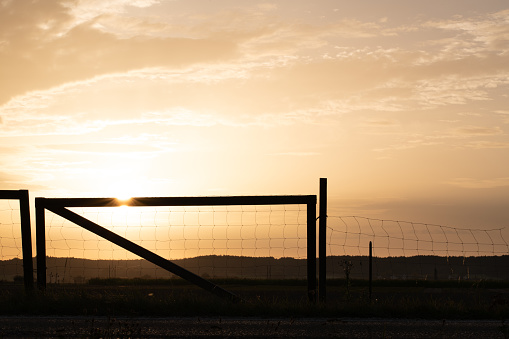The sun sets behind the horizon. An old fence made of wood and wire stands at the side of the road. Fields can be seen.