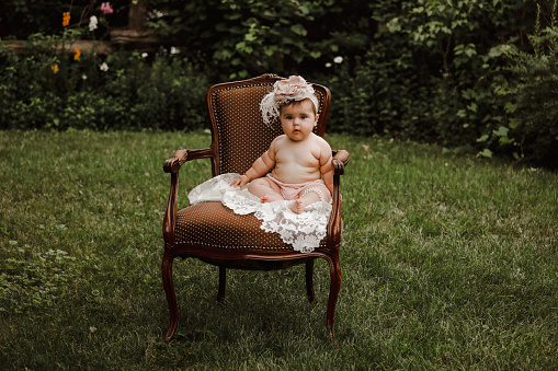 A beautiful baby girl, seven months old, sitting in a vintage chair in a garden.