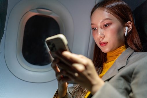 Close Up Portrait of Young Asian Woman on Plane, Engrossed in Music on Smartphone during the Journey. Digital Lifestyles. Connectivity on the move. Business on the go concept.