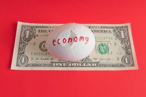 The cracked economy and the value of a dollar bill. A concept image of economic problems represented by a cracked egg over a U.S. dollar bill on red background.