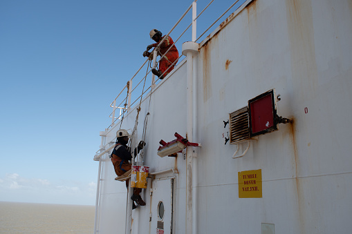 sao luis anchorage, on board ship - november 14, 2023 : crew members of a cargo ship painting on the accomodation bulkhead sitting on a bosun's chair arrangement