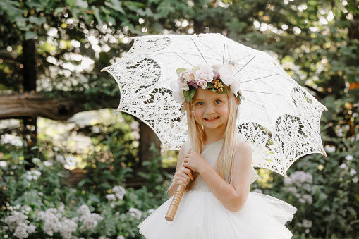 A beautiful little girl, four years old, smiling at the camera as she stands  in a garden under a vintage parasol.