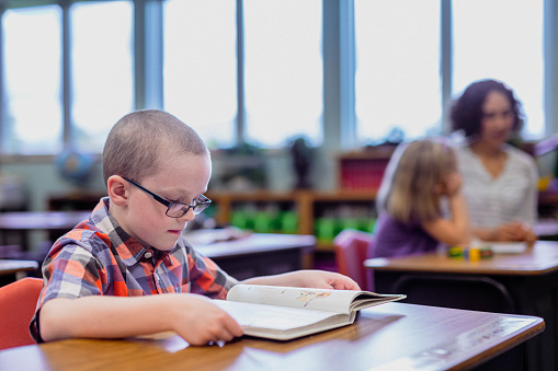 An elementary school age boy wearing glasses sits at a classroom desk, quietly reading a book by himself during a school day.