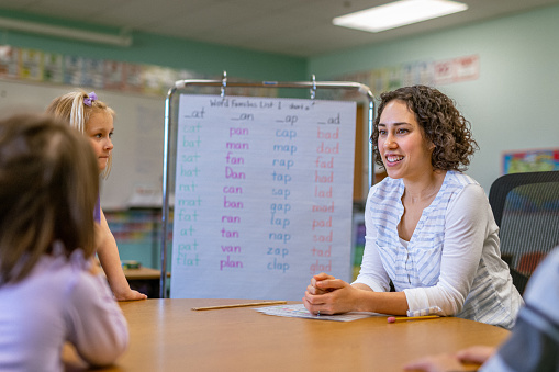 A young female teacher of Eurasian ethnicity sits at a classroom table, teaching a literacy lesson to elementary school students.
