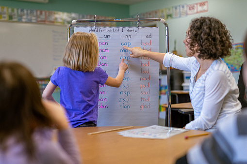 A young teacher of Eurasian ethnicity points to a lesson board as a student stands, learning in an elementary school classroom.