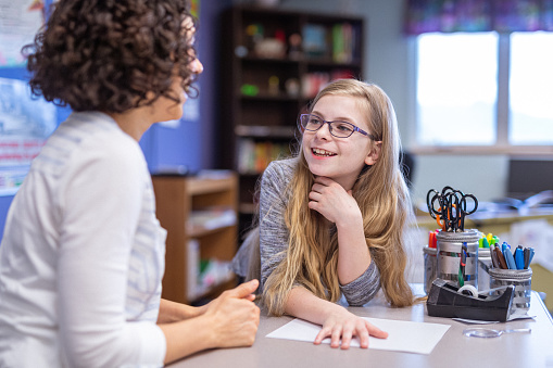 A young girl wearing glasses sits at a classroom table, discussing classwork with her Eurasian teacher during the school day.