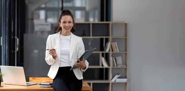 Young happy businesswoman holding pen and file folder while standing  in the office and looking to camera. Copy space.