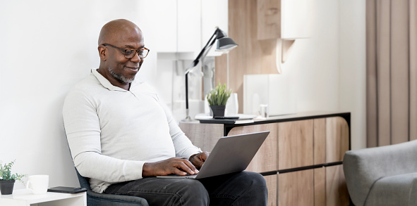 Smiling African American man sitting in living room at home, using laptop. Happy African American Senior Man working from home,