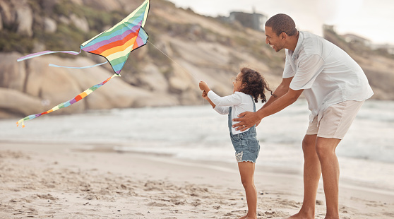 Father teaching child to fly a kite on beach wind with support, love and care. Helping, learning and fun outdoor with dad and girl kid together on holiday or summer vacation by ocean water and sand