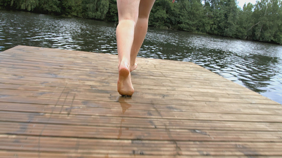 Legs of a young woman walking on the pier. A woman walks along a pier on the river on a summer day. Rest on the river in the village