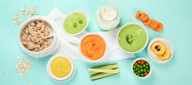 Infant baby food. Bowls with vegetable fruit puree, green, orange, yellow colors - broccoli, carrots, banana, apple on blue background. Top view. Panorama.