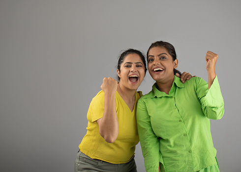 Portrait of ecstatic successful female friends shaking fists and standing cheerfully against gray background
