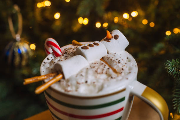 Christmas hot chocolate mug with snowman made of marshmallows and candy cane over Xmas tree background