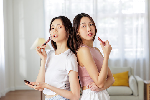 Two Asian women apply makeup meticulously, focusing on perfecting their skin care and beauty routine in a well-lit room. Skin Care Concept