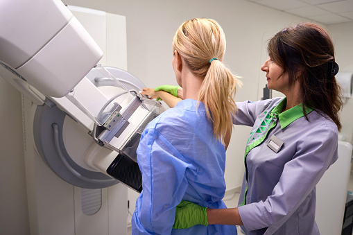 Adult female patient reaching for handgrip on mammography unit assisted by experienced mammographer