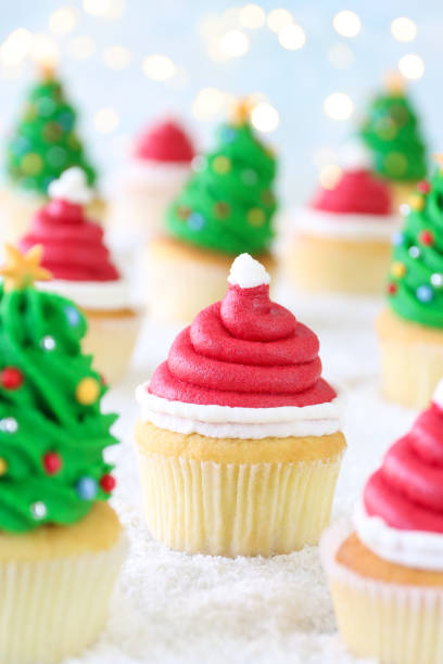 istock Close-up image batch homemade, Santa Claus hat and Christmas tree design cupcakes, red buttercream swirl with white icing bobbles, green buttercream swirl with multicoloured sugar sprinkles, icing sugar snow, illuminated fairy lights background 1799976593