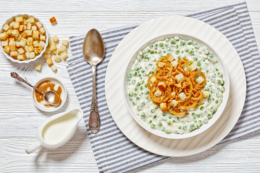 creamed green peas topped with french fried onions and croutons in white bowl on white wooden table with spoon and ingredients, horizontal view from above, flat lay