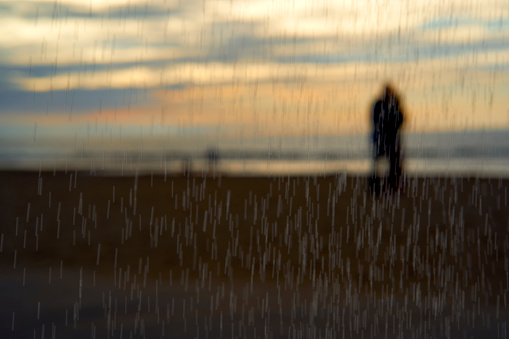 Out of focus. Lovers also couple on sandy beach looking into the sunset. Raindrops fall from the sky. In silhouette.