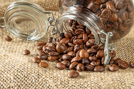 Coffee theme. Coffee beans pour out of a glass jar onto a table covered with burlap.