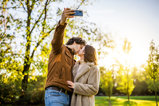 Portrait of happy loving couple in park in sunset. They are kissing and taking selfie.