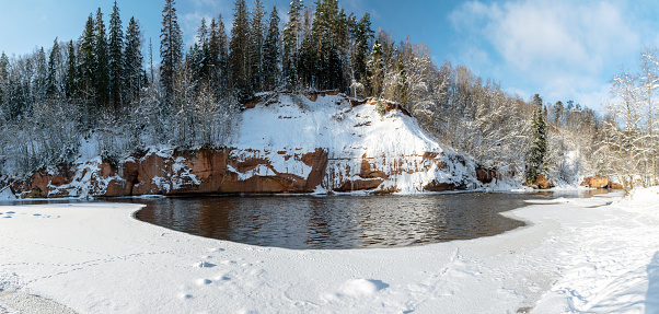 panoramic winter landscape with snowy sandstone cliff and frozen ice falls, slow river water, snowy trees on the river bank, Kuku cliffs, river Gauja, Latvia