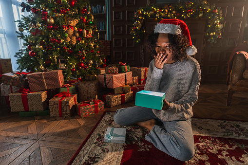 Merry Christmas. African American woman unpacking open up gift box near Christmas tree. Girl in living room with Christmas tree and fireplace opening gift box with surprise face. Christmas eve at home