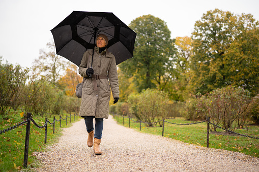 one mid adult woman wearing warm coat and umbrella walking alone on path in public park on rainy day in autumn, low angle view