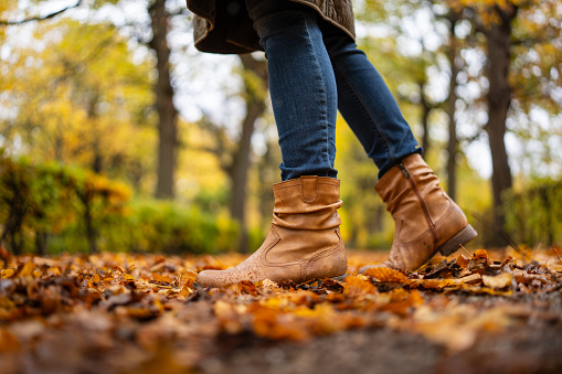 side view of walking boots, detail of womans legs wearing wet shoes because of rain on cold autumn day outdoors in park alley covered in autumn leaves, low angle view with shallow focus