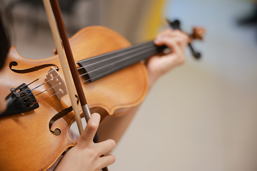 A smiling little girl sits next to her attentive music teacher and plays the violin during music class. Her teacher adjusts the violin's position as she plays.