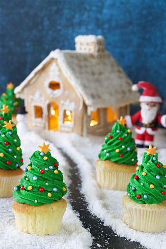 Stock photo showing close-up view of a batch of freshly baked, homemade Christmas tree design cupcakes, in paper cake cases, displayed with a gingerbread house in a snowy, Christmas scene. The cupcakes are topped with swirls of green butter icing and decorated with multicoloured sugar sprinkles. Home baking concept.
