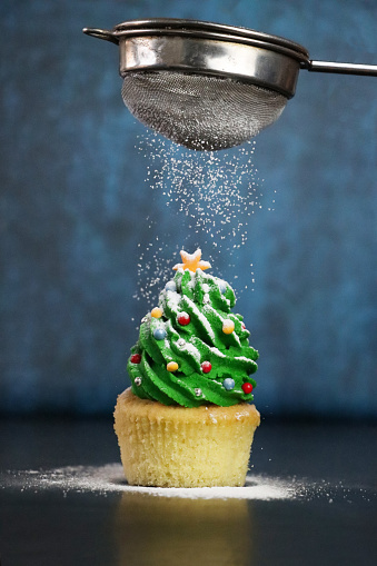 Stock photo showing close-up view of a freshly baked, homemade Christmas tree design cupcake displayed on icing sugar snow against a blue mottled background. The cake is topped with a swirl of green butter icing and decorated with multicoloured sugar sprinkles. Home baking concept.