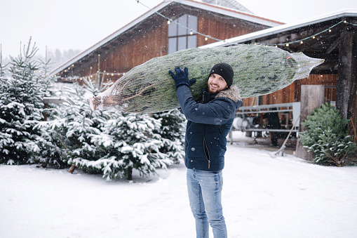 Handsome bearded man holding wrapped Christmas tree on fair. Happy male bring fir tree.