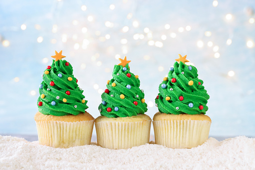 Stock photo showing close-up view of a row of freshly baked, homemade Christmas tree design cupcakes, in paper cake cases, displayed surrounded by illuminated fairy lights on icing sugar snow against a pale blue background. The cakes are topped with swirls of green butter icing and decorated with multicoloured sugar sprinkles. Home baking concept.